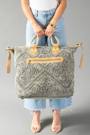 A person in jeans and sandals holds the Gray Vintage Inspired Textured Tote by Panache Apparel Collection, featuring tan suede handles and a zipper closure, against a plain gray background.