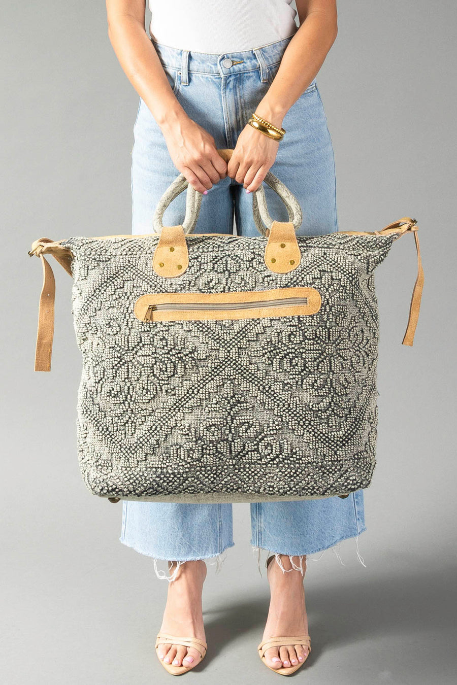 A person in jeans and sandals holds the Gray Vintage Inspired Textured Tote by Panache Apparel Collection, featuring tan suede handles and a zipper closure, against a plain gray background.