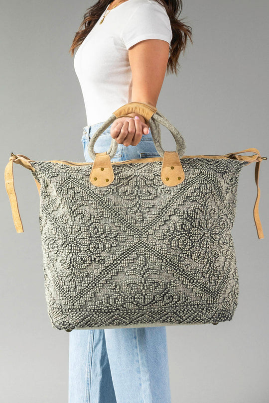 A woman in a white shirt and blue jeans holds the Gray Vintage Inspired Textured Tote by Panache Apparel Collection, featuring suede leather handles and a geometric design, against a plain gray background.