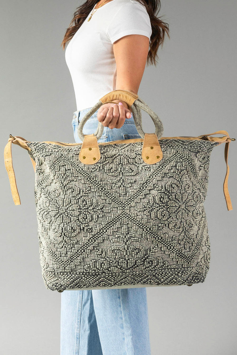 A woman in a white shirt and blue jeans holds the Gray Vintage Inspired Textured Tote by Panache Apparel Collection, featuring suede leather handles and a geometric design, against a plain gray background.