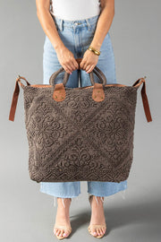 A person dressed in a white top and light blue jeans holds the Panache Apparel Collection’s Brown Vintage Inspired Textured Tote, featuring leather handles and side straps. The individual accessorizes with sandals and a bracelet, set against a plain gray background.