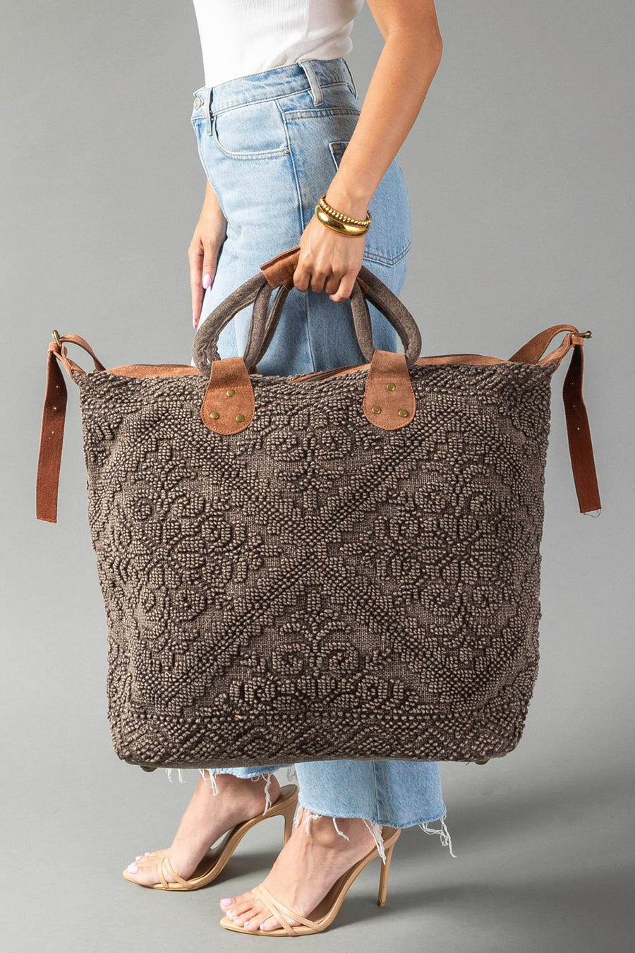 A person in jeans and heels holds the Panache Apparel Collection’s Brown Vintage Inspired Textured Tote, a large bag with short handles, side straps, an intricate woven pattern, and brown leather accents.