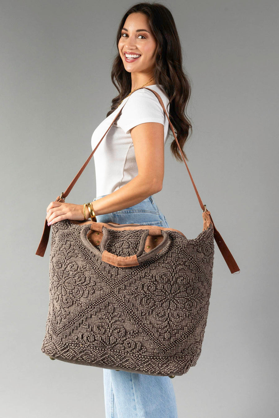 A woman in a white top and blue jeans smiles while carrying the Panache Apparel Collection Brown Vintage Inspired Textured Tote, featuring leather handles and a shoulder strap, against a plain gray background.