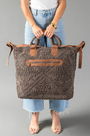 A person wearing a white top and blue jeans holds the Panache Apparel Collection Brown Vintage Inspired Textured Tote, which features leather handles and a front zipper pocket. The tote rests against their legs on a gray background.