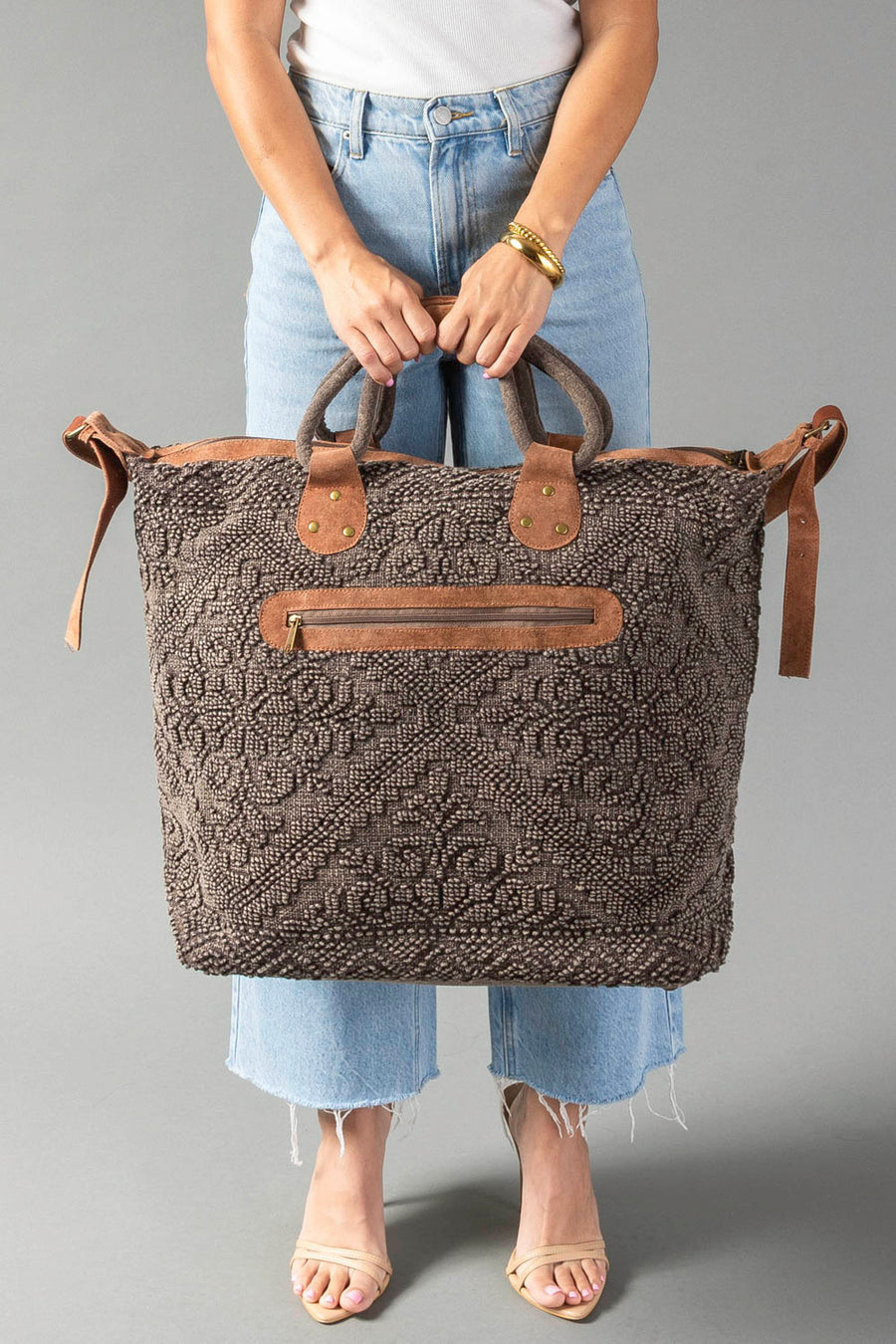 A person wearing a white top and blue jeans holds the Panache Apparel Collection Brown Vintage Inspired Textured Tote, which features leather handles and a front zipper pocket. The tote rests against their legs on a gray background.