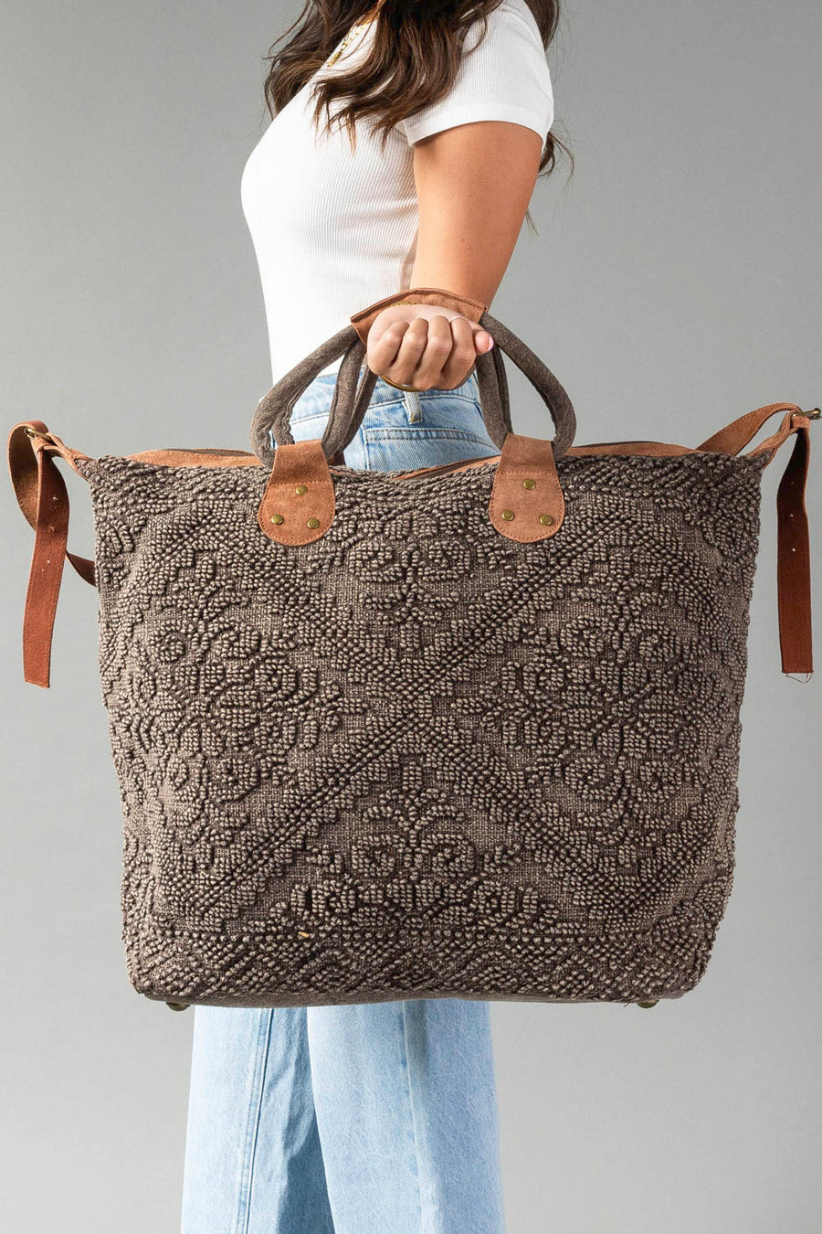 A woman in a white top and jeans holds the Panache Apparel Collection Brown Vintage Inspired Textured Tote, featuring leather handles and decorative stitching, against a plain gray background.