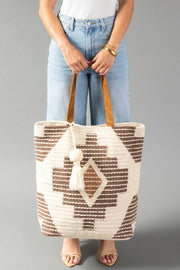 A person in a white top and blue jeans holds the Cream, Brown & Rust Western Tote from Panache Apparel Collection, featuring brown geometric patterns, tan handles, and a cream tassel. The background is plain gray.