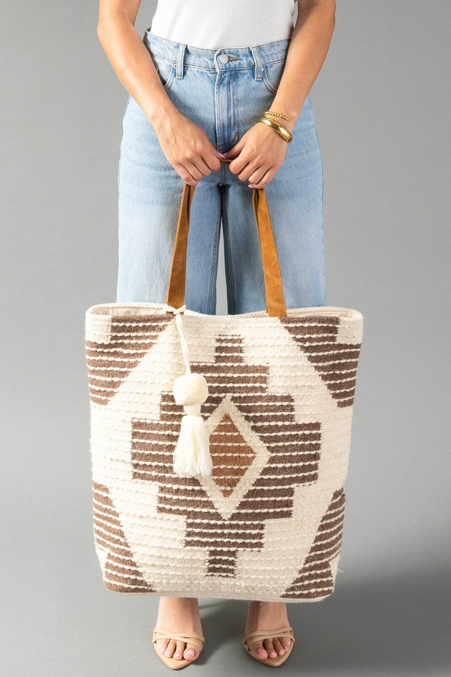 A person in a white top and blue jeans holds the Cream, Brown & Rust Western Tote from Panache Apparel Collection, featuring brown geometric patterns, tan handles, and a cream tassel. The background is plain gray.