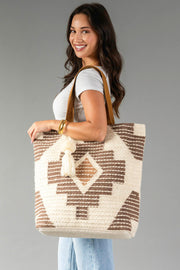 A woman smiles against a plain gray background, wearing a white top and jeans while holding the Panache Apparel Collection Cream, Brown & Rust Western Tote, a large bag featuring a beige and brown pattern, leather straps, and a tassel.