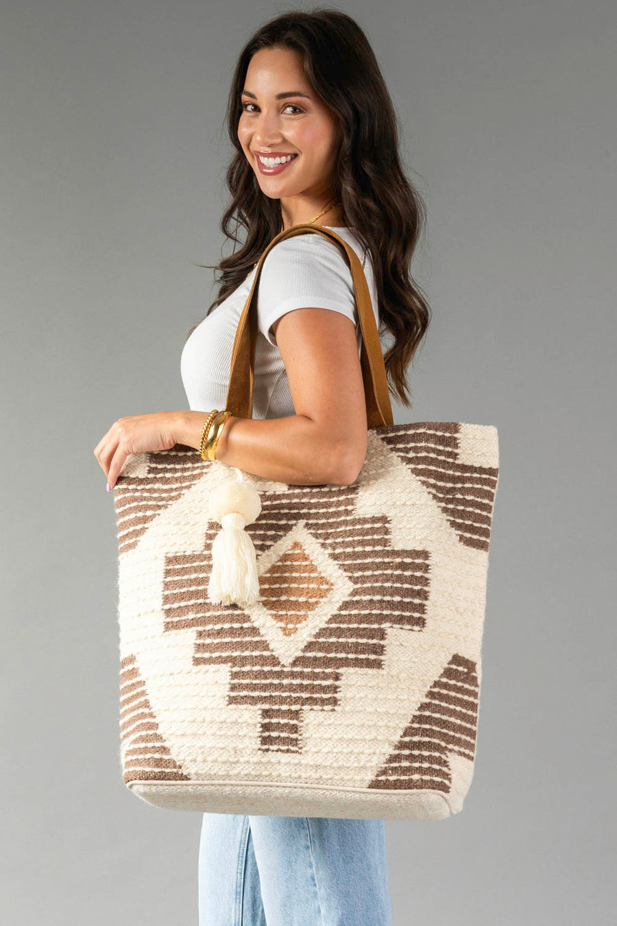 A woman smiles against a plain gray background, wearing a white top and jeans while holding the Panache Apparel Collection Cream, Brown & Rust Western Tote, a large bag featuring a beige and brown pattern, leather straps, and a tassel.