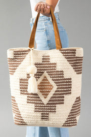 A person in jeans holds the Cream, Brown & Rust Western Tote by Panache Apparel Collection, featuring brown geometric patterns, tan handles, and a white tassel, against a gray background.