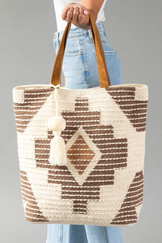 A person in jeans holds the Cream, Brown & Rust Western Tote by Panache Apparel Collection, featuring brown geometric patterns, tan handles, and a white tassel, against a gray background.