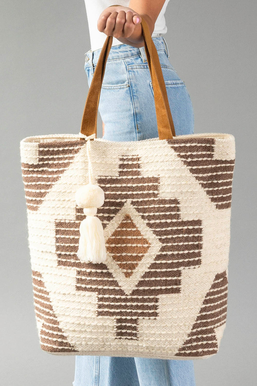 A person in jeans holds the Cream, Brown & Rust Western Tote by Panache Apparel Collection, featuring brown geometric patterns, tan handles, and a white tassel, against a gray background.
