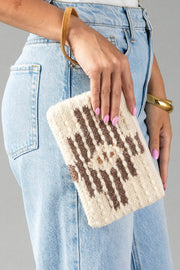 A person wearing light blue jeans holds the Panache Apparel Collection’s Cream, Brown & Rust Western Wristlet, which features a tan wrist strap and a woven pattern. Their nails are painted pink and they wear a gold bracelet.