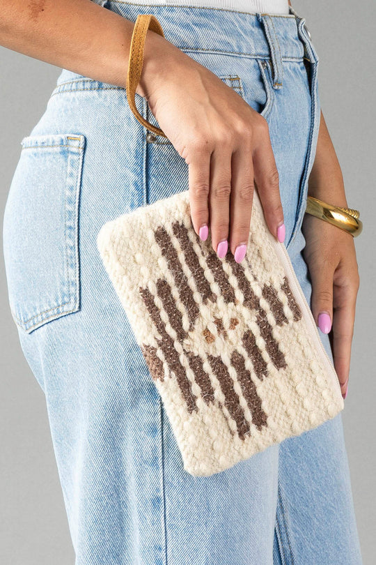 A person wearing light blue jeans holds the Panache Apparel Collection’s Cream, Brown & Rust Western Wristlet, which features a tan wrist strap and a woven pattern. Their nails are painted pink and they wear a gold bracelet.