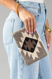 A person with light pink nail polish holds the Panache Apparel Collection’s Taupe, Cream, Brown & Black Western Wristlet, which features a geometric pattern, while standing next to light blue jeans.