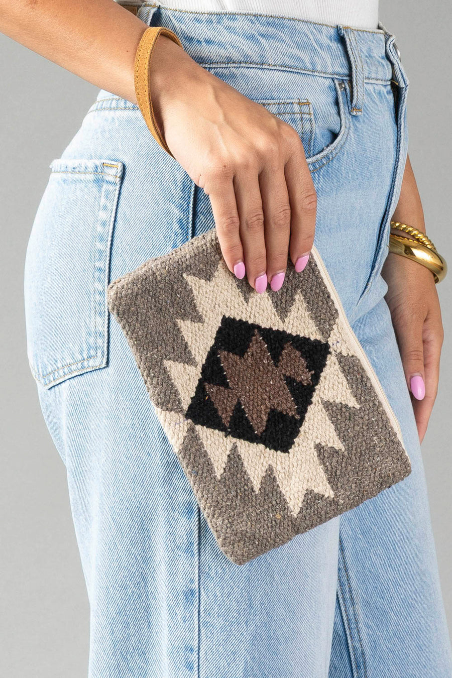 A person with light pink nail polish holds the Panache Apparel Collection’s Taupe, Cream, Brown & Black Western Wristlet, which features a geometric pattern, while standing next to light blue jeans.