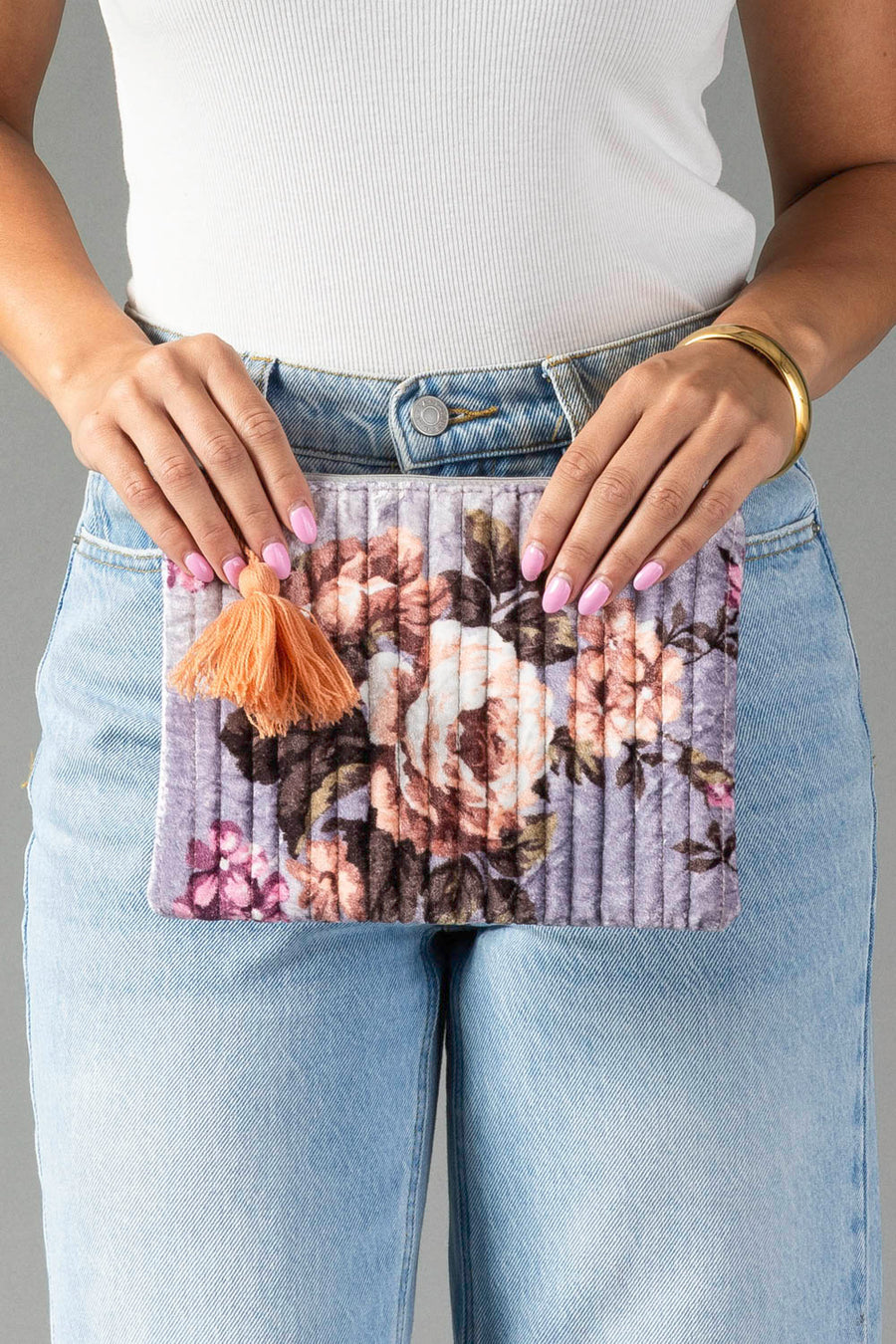 A person in a white top and blue jeans holds the Lavender & Multicolored Floral Pouch from Panache Apparel Collection, featuring a quilted floral design and a peach tassel. Their nails are painted light pink and they wear a gold bracelet.