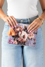A woman in a white top and blue jeans holds the Panache Apparel Collection’s Lavender & Multicolored Floral Pouch, adorned with a peach tassel. Her nails are painted light pink, and she wears a gold bracelet.