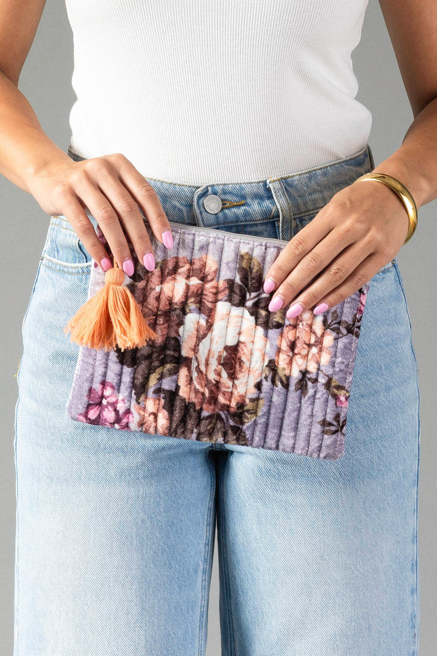 A woman in a white top and blue jeans holds the Panache Apparel Collection’s Lavender & Multicolored Floral Pouch, adorned with a peach tassel. Her nails are painted light pink, and she wears a gold bracelet.