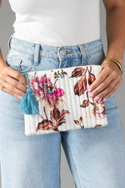 A person in a white top and blue jeans holds the Panache Apparel Collection White & Multicolored Floral Pouch, featuring pink and brown flowers, vertical quilting, and a blue tassel.
