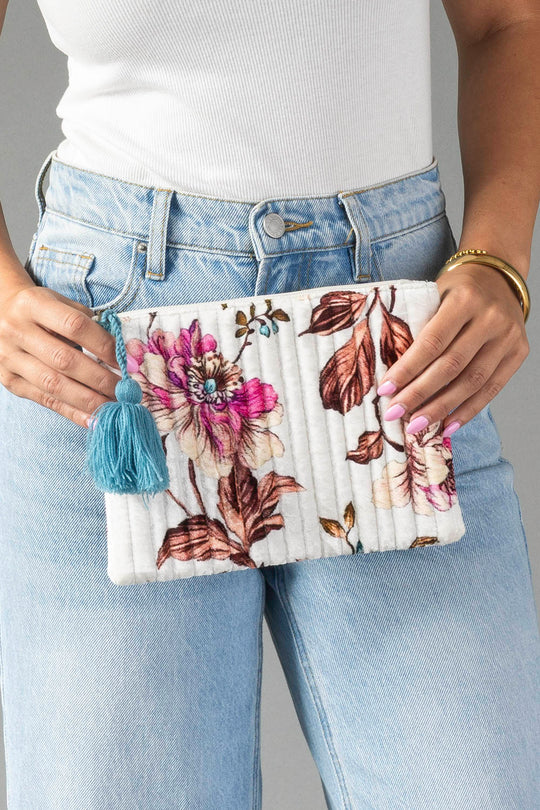 A person in a white top and blue jeans holds the Panache Apparel Collection White & Multicolored Floral Pouch, featuring pink and brown flowers, vertical quilting, and a blue tassel.