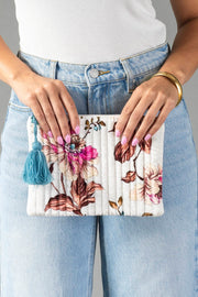 A person in blue jeans and a white top holds the Panache Apparel Collection White & Multicolored Floral Pouch, featuring a blue tassel. Their nails are painted pastel pink, and they wear a gold bangle on one wrist.