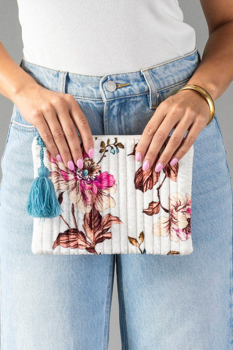 A person in blue jeans and a white top holds the Panache Apparel Collection White & Multicolored Floral Pouch, featuring a blue tassel. Their nails are painted pastel pink, and they wear a gold bangle on one wrist.