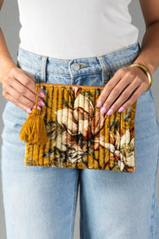A person dressed in a white top and blue jeans holds the Mustard & Multicolored Floral Pouch from Panache Apparel Collection, featuring a quilted design with a yellow tassel. Their nails are painted lavender, and a gold bangle adorns one wrist.