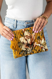 A person in light jeans and a white top holds the Panache Apparel Collection Mustard & Multicolored Floral Pouch, which features a tassel detail. The person has light purple nail polish and wears a gold bangle.