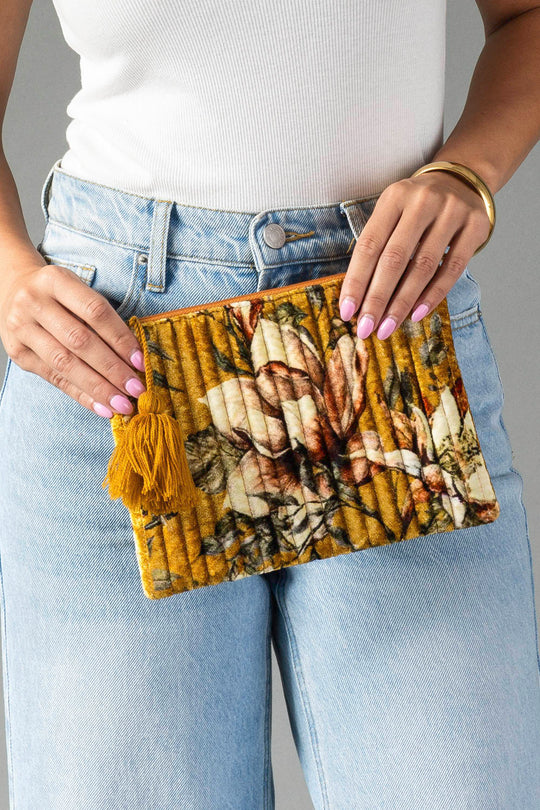 A person in light jeans and a white top holds the Panache Apparel Collection Mustard & Multicolored Floral Pouch, which features a tassel detail. The person has light purple nail polish and wears a gold bangle.