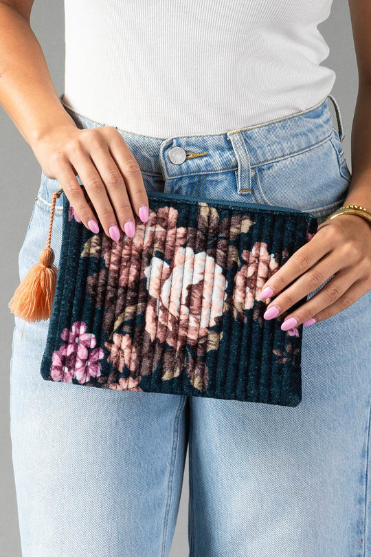 A person in a white top and light blue jeans holds the Panache Apparel Collection’s Navy & Multicolored Floral Pouch, a small rectangular clutch featuring a tassel. Their nails are painted light pink and they wear a simple bracelet on one wrist.
