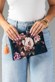 A person in jeans and a white top holds the Panache Apparel Collection’s Navy & Multicolored Floral Pouch, featuring a peach tassel. Their nails are painted lilac, and they wear a gold bangle.