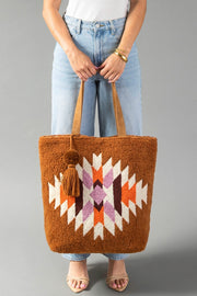A person in jeans and a white top holds the Orange, Cream & Pink Western Tote by Panache Apparel Collection, a large tote bag featuring a geometric pattern in cream, orange, and pink. The bag is finished with tan handles and a decorative pom-pom tassel accessory.