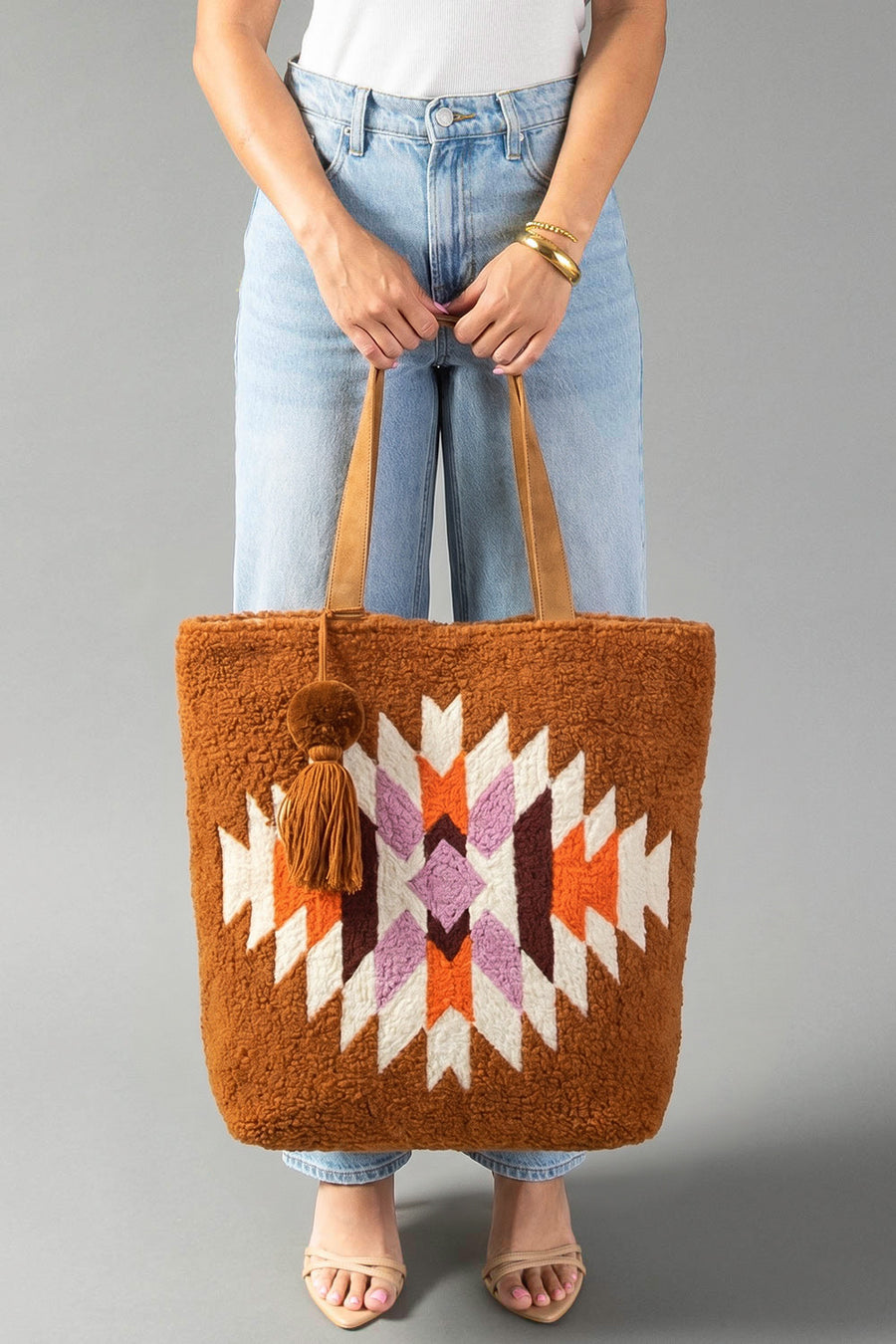 A person in jeans and a white top holds the Orange, Cream & Pink Western Tote by Panache Apparel Collection, a large tote bag featuring a geometric pattern in cream, orange, and pink. The bag is finished with tan handles and a decorative pom-pom tassel accessory.