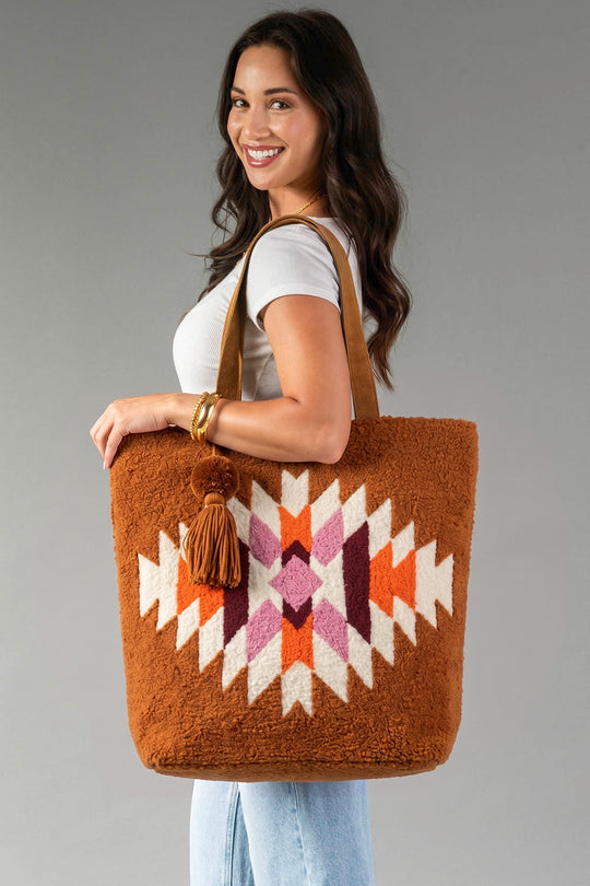 A woman in a white shirt and jeans smiles while holding the Panache Apparel Collection Orange, Cream & Pink Western Tote, featuring a colorful geometric pattern and a pom-pom accent, against a gray background.