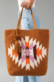 A person holds the Orange, Cream & Pink Western Tote by Panache Apparel Collection, a large brown bag featuring leather handles, a geometric pattern in white, orange, purple, and black, and adorned with a tassel and pompom accessory.