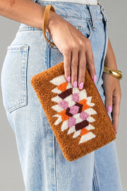 A person wearing light blue jeans holds the Orange, Cream & Pink Western Wristlet from Panache Apparel Collection, a brown textured clutch featuring colorful geometric patterns in white, orange, and purple. Their nails are painted pink and they accessorize with a gold bracelet.