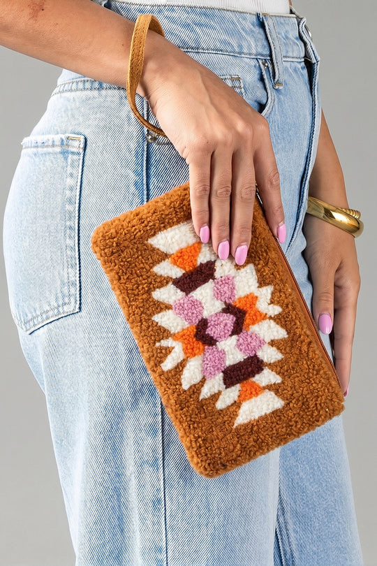 A person wearing light blue jeans holds the Orange, Cream & Pink Western Wristlet from Panache Apparel Collection, a brown textured clutch featuring colorful geometric patterns in white, orange, and purple. Their nails are painted pink and they accessorize with a gold bracelet.