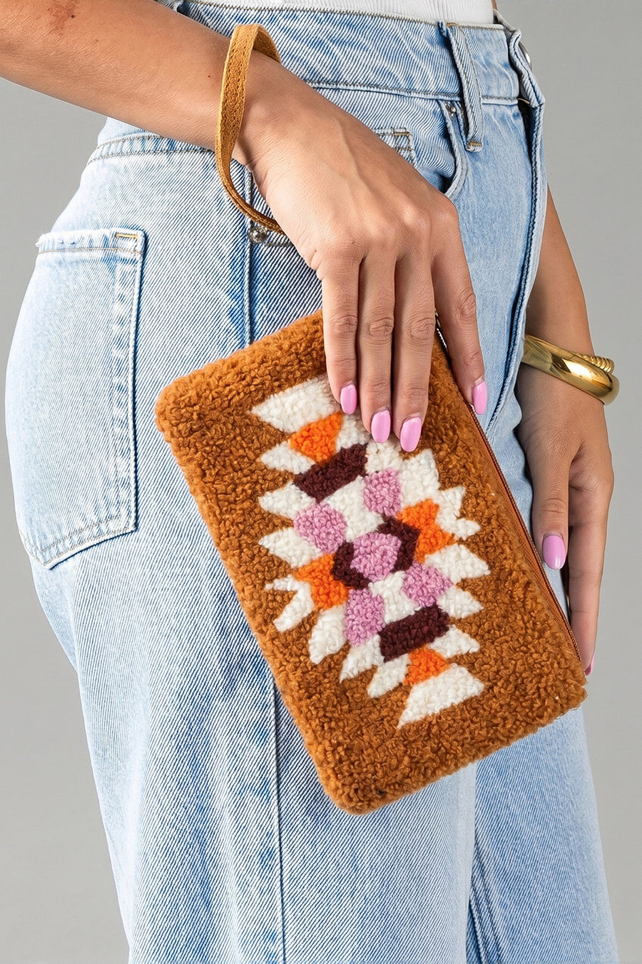 A person wearing light blue jeans holds the Orange, Cream & Pink Western Wristlet from Panache Apparel Collection, a brown textured clutch featuring colorful geometric patterns in white, orange, and purple. Their nails are painted pink and they accessorize with a gold bracelet.