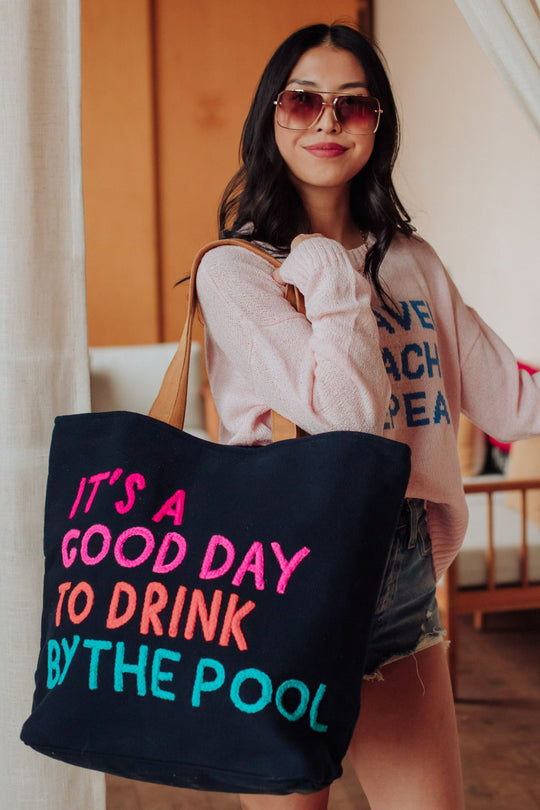 A woman in sunglasses, a light pink sweater, and denim shorts smiles while holding the Panache Apparel Collection Navy Drink By The Pool Tote, featuring colorful "ITS A GOOD DAY TO DRINK BY THE POOL" text.