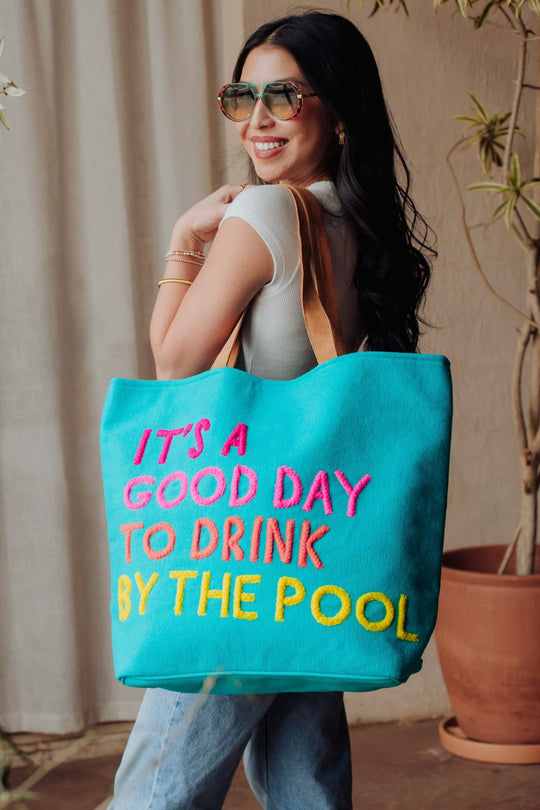 A smiling woman in sunglasses and a white top carries the Blue Drink By The Pool Tote by Panache Apparel Collection, featuring colorful "ITS A GOOD DAY TO DRINK BY THE POOL" text, while standing near potted plants.