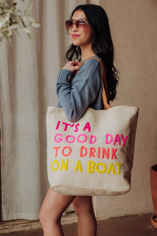 A woman with long dark hair, sunglasses, and a blue sweater smiles while holding the Panache Apparel Collection Cream Drink On A Boat Tote featuring colorful “IT’S A GOOD DAY TO DRINK ON A BOAT” text and leather suede straps.