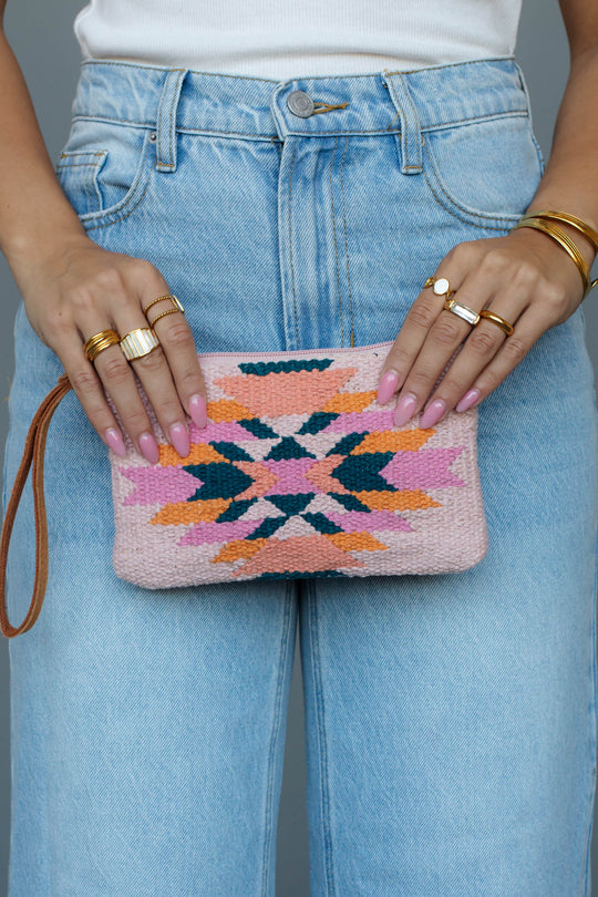 A person wearing a white top and blue jeans holds the panacheaccessories Pink, Teal & Orange Western Wristlet, a small pink woven clutch with a colorful geometric pattern, western-inspired wristlet, and leather pull. They wear gold rings, bangles, and pink nail polish.