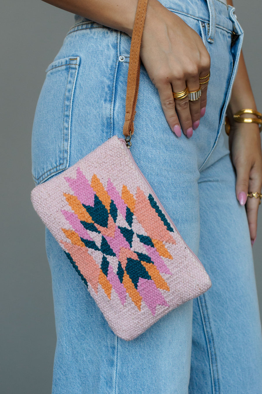 A person in light-wash jeans holds the panacheaccessories Pink, Teal & Orange Western Wristlet—a woven clutch with a geometric pattern and tan strap—showing pink nails and gold jewelry. The bag is cotton-lined.