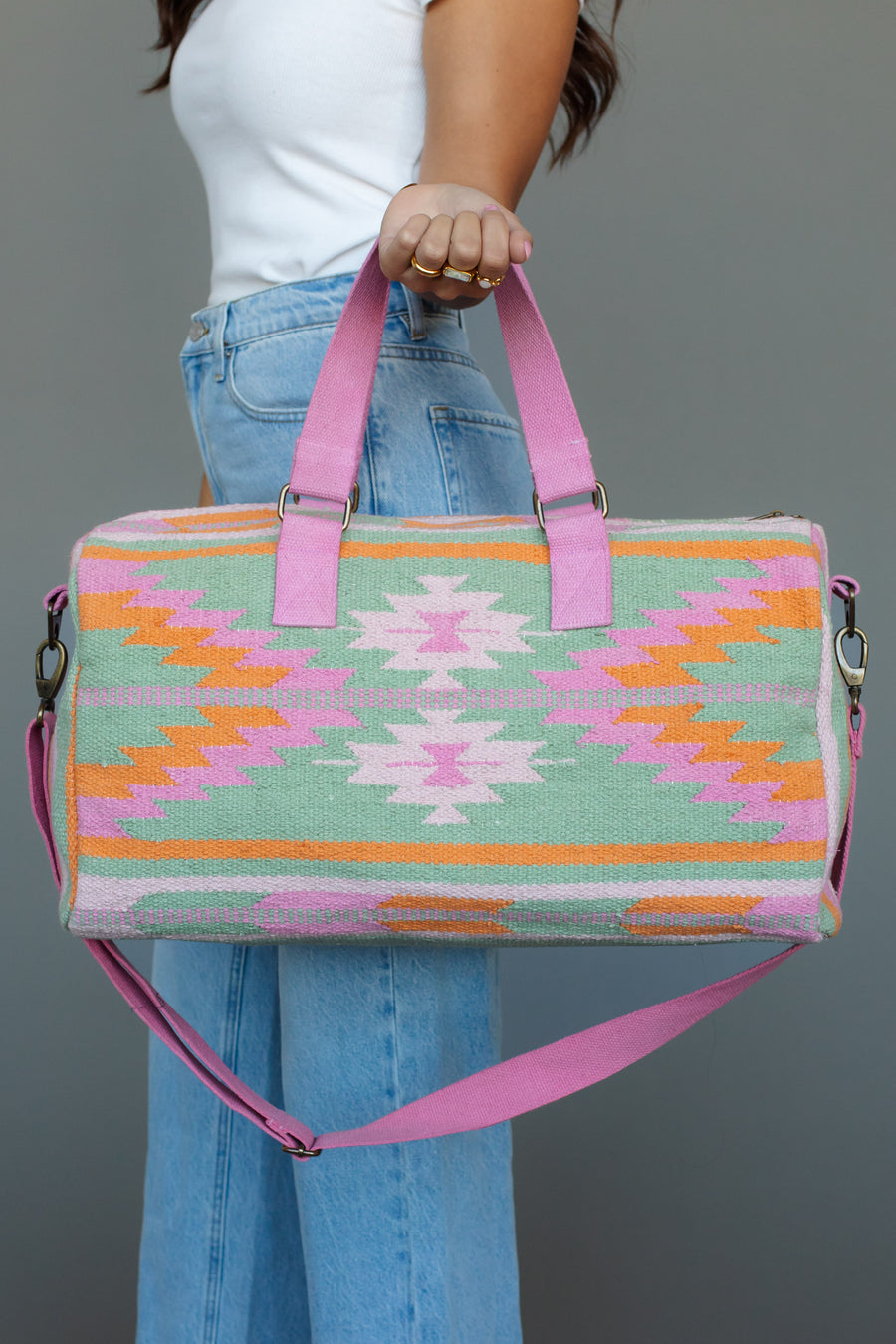 A woman in a white top and blue jeans holds the Mint, Pink & Orange Western Duffel by panacheaccessories, featuring pink handles and vibrant geometric patterns, against a plain gray background.