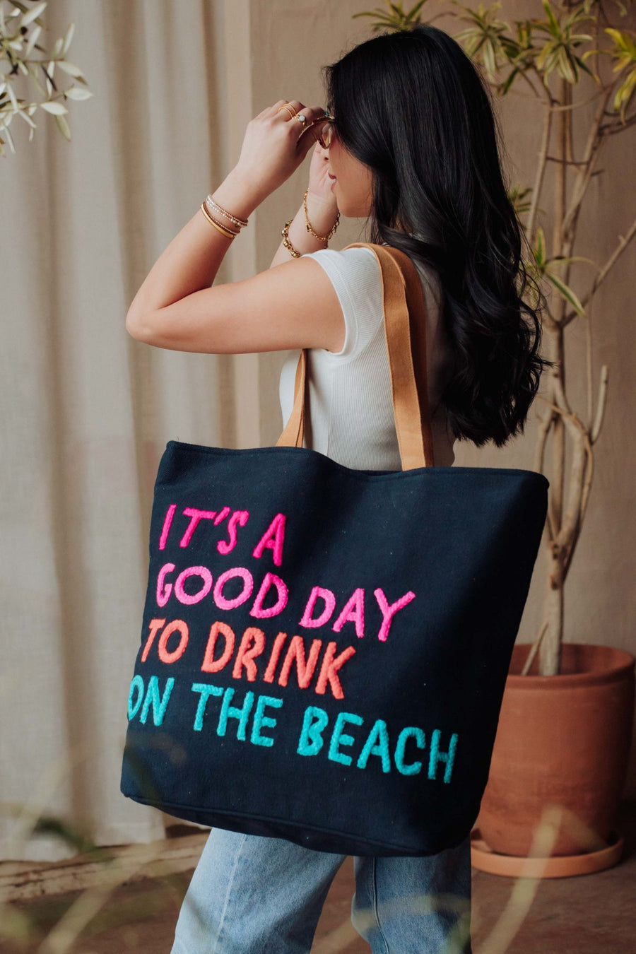 A woman with long dark hair in a white top and blue jeans holds the Panache Apparel Collection Navy Drink On The Beach Tote, featuring the text "ITS A GOOD DAY TO DRINK ON THE BEACH," standing indoors near potted plants.