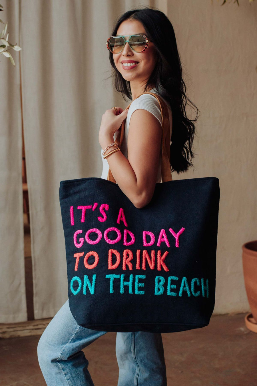 A smiling woman in sunglasses and a white top holds the Navy Drink On The Beach Tote by Panache Apparel Collection, featuring text: “ITS A GOOD DAY TO DRINK ON THE BEACH.” She stands indoors near a beige curtain and potted plant.