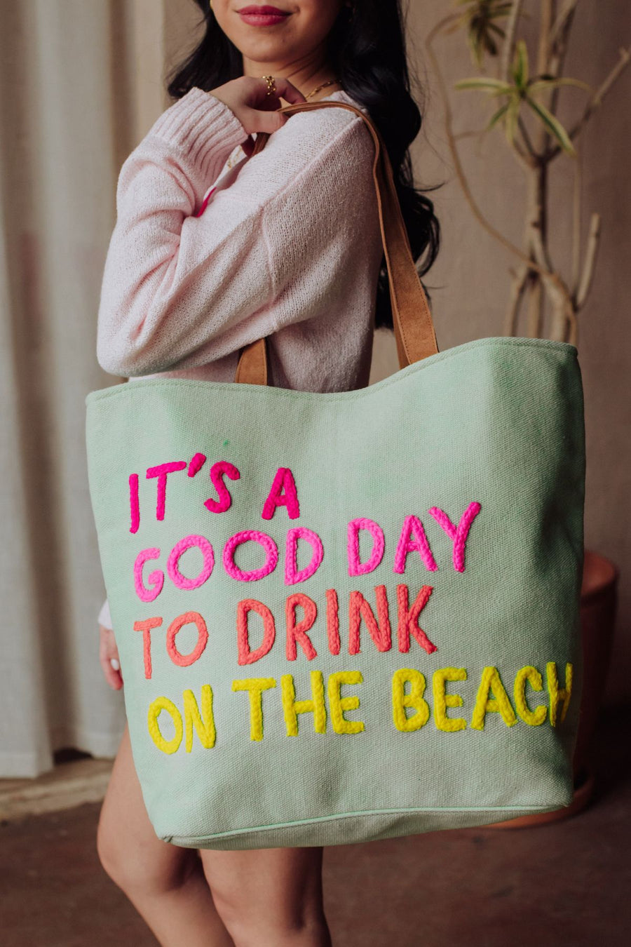A woman wearing a light pink sweater holds the Panache Apparel Collection Mint Drink On The Beach Tote, featuring faux suede straps and colorful embroidered text: "It's a good day to drink on the beach.