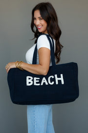 A woman with long dark hair, dressed in a white t-shirt and light blue jeans, smiles while carrying the Panache Apparel Collection Beach Tote—a large navy blue bag with "BEACH" in white—and wears gold bangles on her wrist.